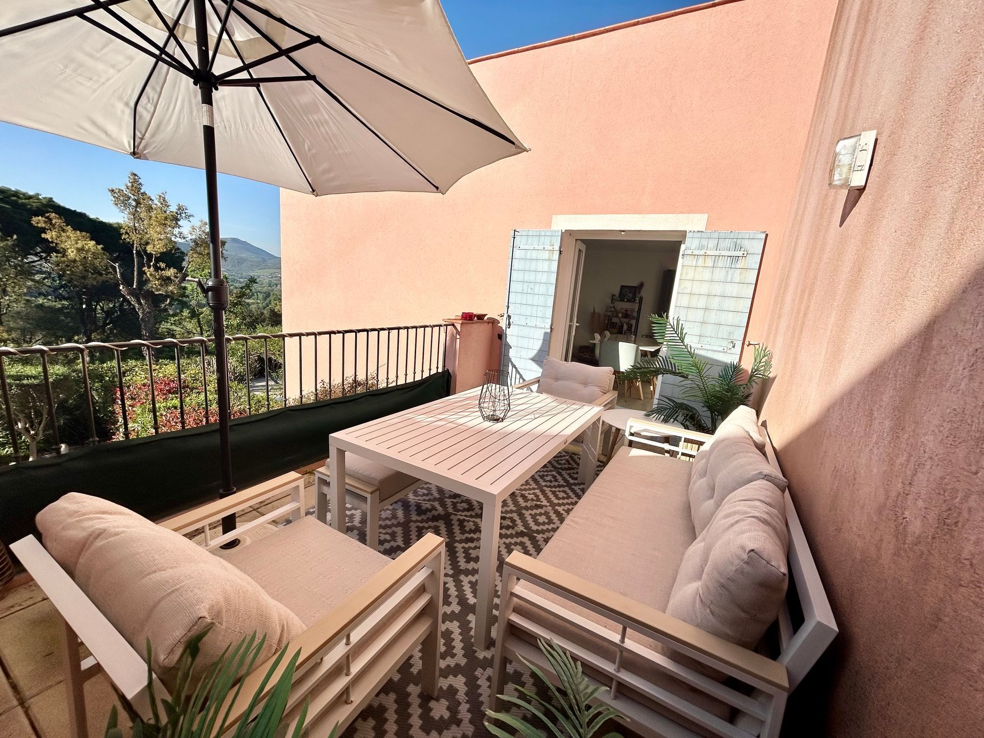Terrasse extérieure avec sièges, table et parasol. Les murs sont roses et offrent une vue sur les arbres et les montagnes.