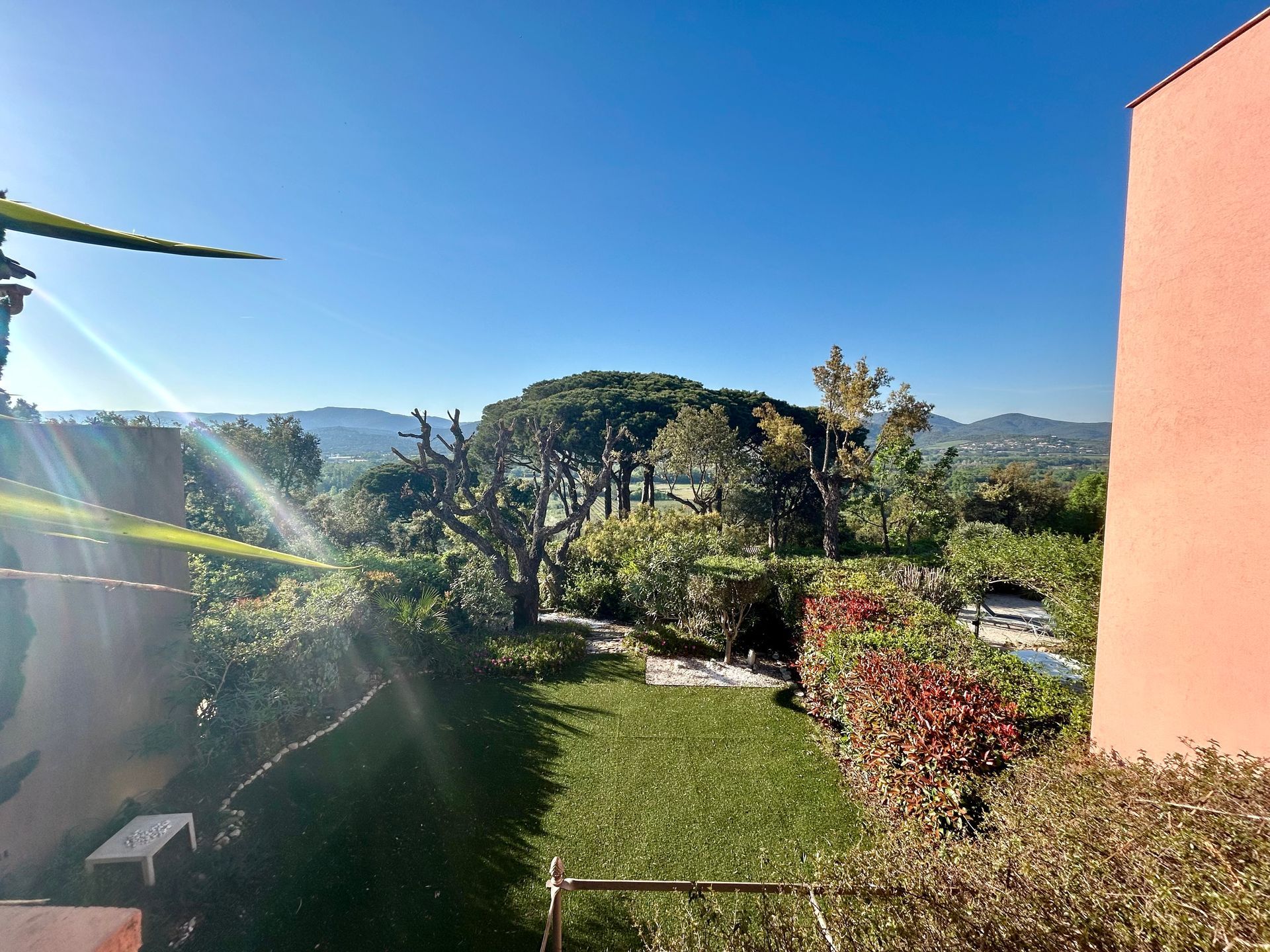 Jardin verdoyant et luxuriant avec des arbres, un ciel bleu ensoleillé et une vue sur les collines lointaines.