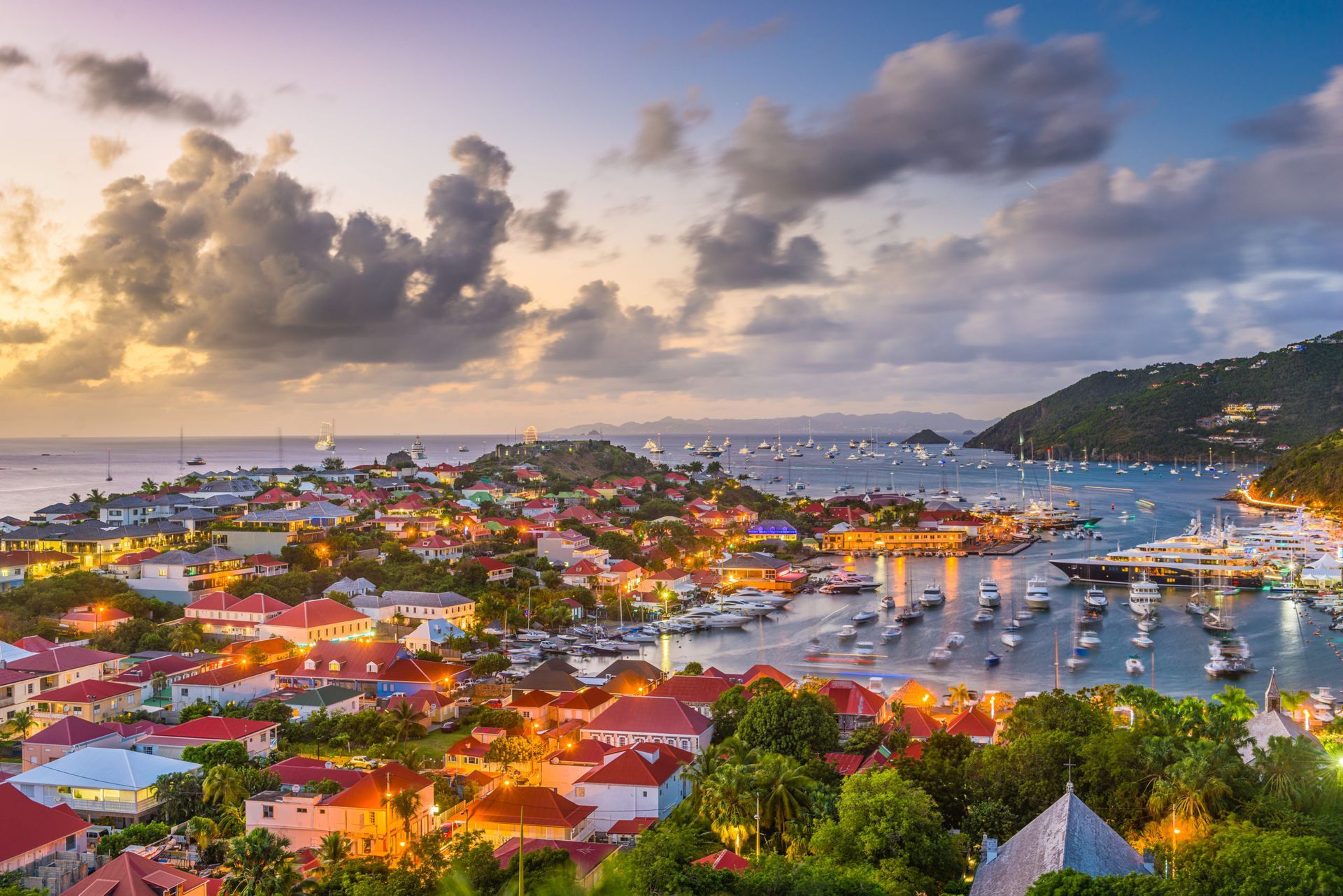 Ville côtière au crépuscule, avec des bâtiments et des bateaux dans le port, sous un ciel nuageux.
