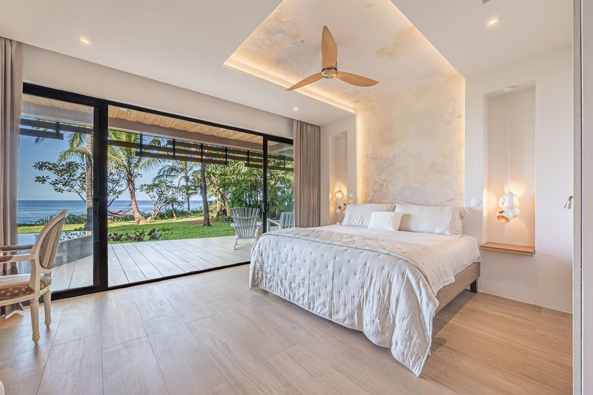 Chambre lumineuse avec vue sur l'océan, portes coulissantes en verre, literie blanche, tons neutres.