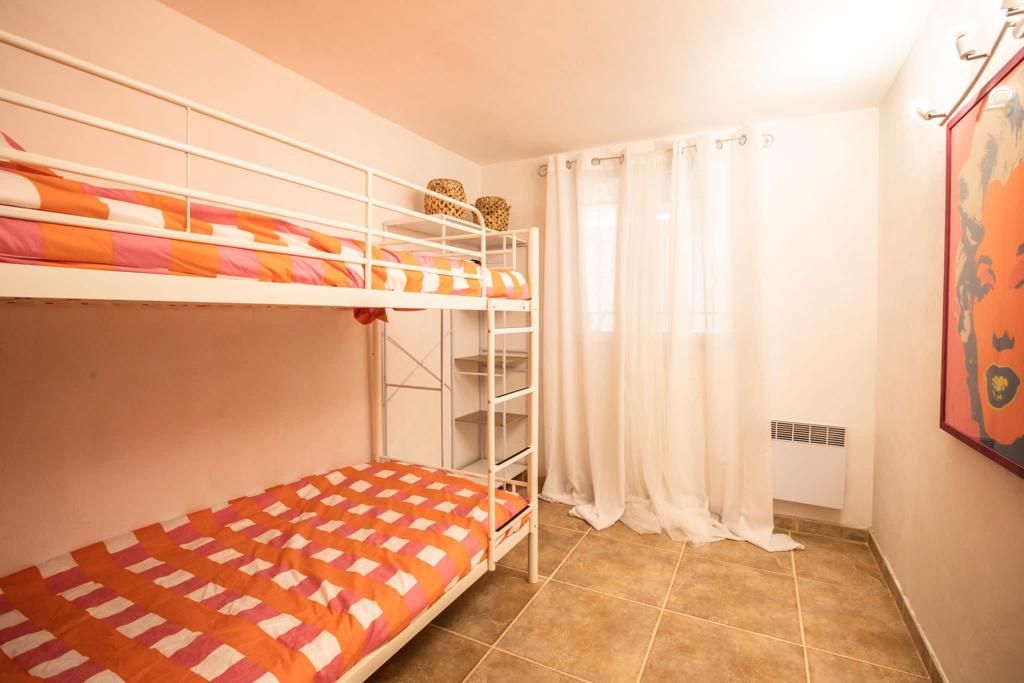 Lits superposés dans une chambre, literie orange et blanche, murs blancs, fenêtre à rideaux, tableau au mur, sol carrelé.