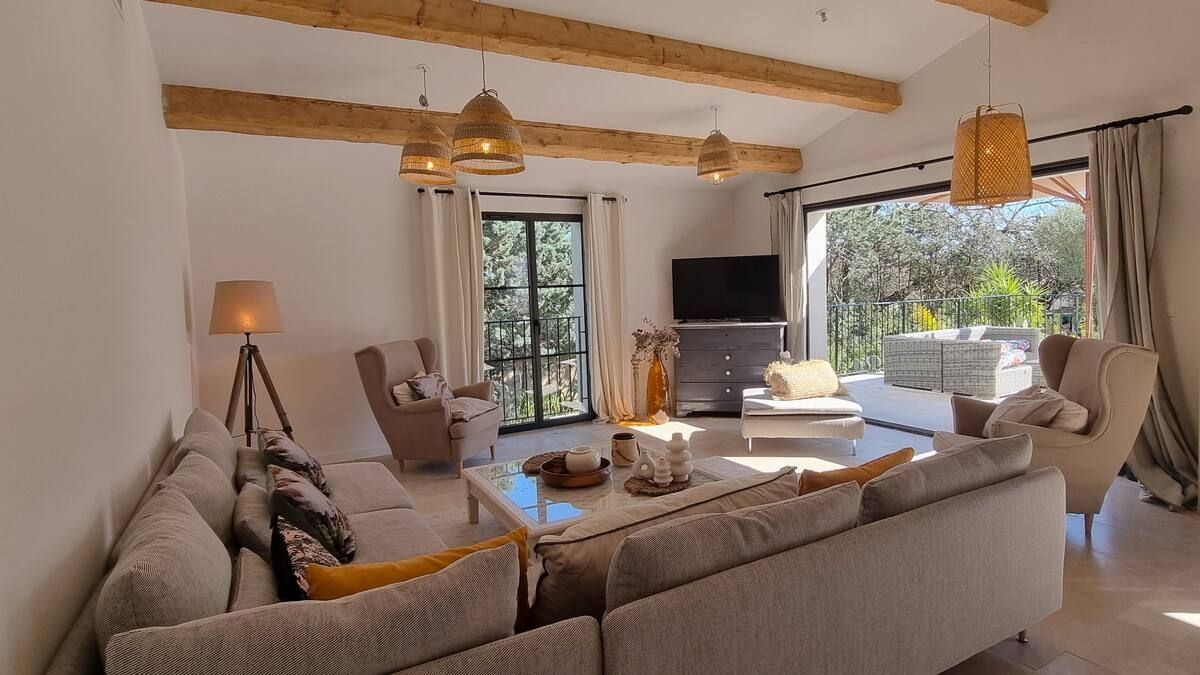 Séjour lumineux avec canapé beige, poutres en bois et grandes fenêtres donnant sur la verdure.