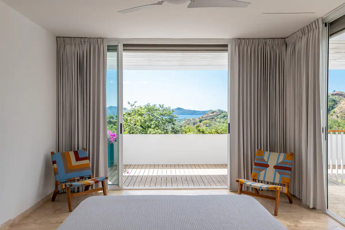 Chambre avec portes ouvertes, balcon avec vue sur les arbres et l'océan. Deux chaises encadrent le lit.