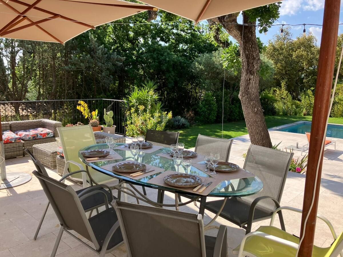 Table à manger d'extérieur aménagée pour un repas, sous un grand parasol, avec une piscine en arrière-plan.