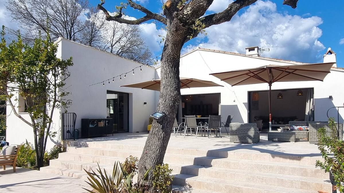 Maison blanche avec terrasse, parasols et marches en pierre sous un ciel bleu et un arbre.
