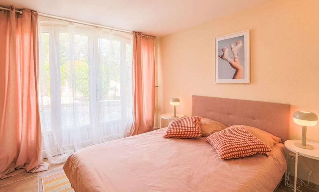 Chambre avec murs et linge de lit roses ; grande fenêtre avec rideaux, lit avec oreillers, deux tables de chevet.