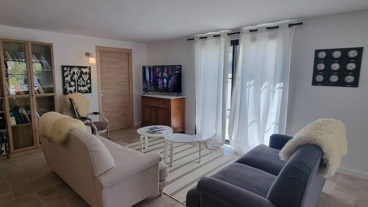 Salon avec deux canapés, petite table, télévision, tapis, rideaux transparents et porte en bois clair.