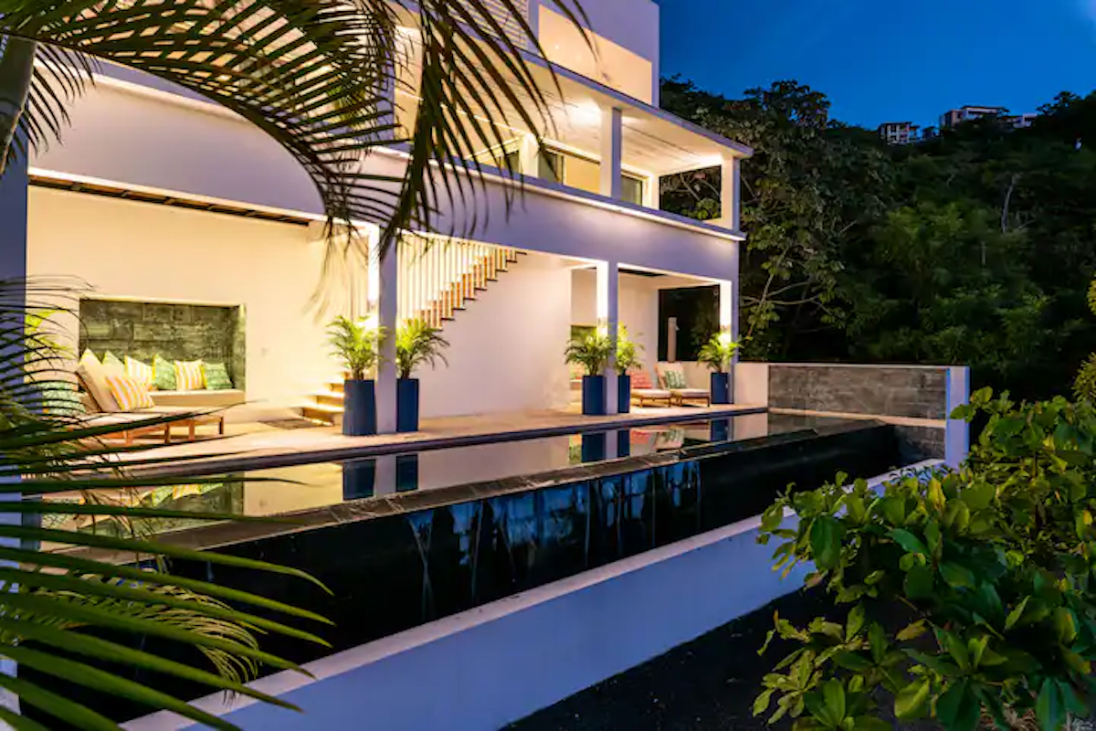 Villa blanche avec piscine au crépuscule ; palmier, plantes, escaliers et colline en arrière-plan.
