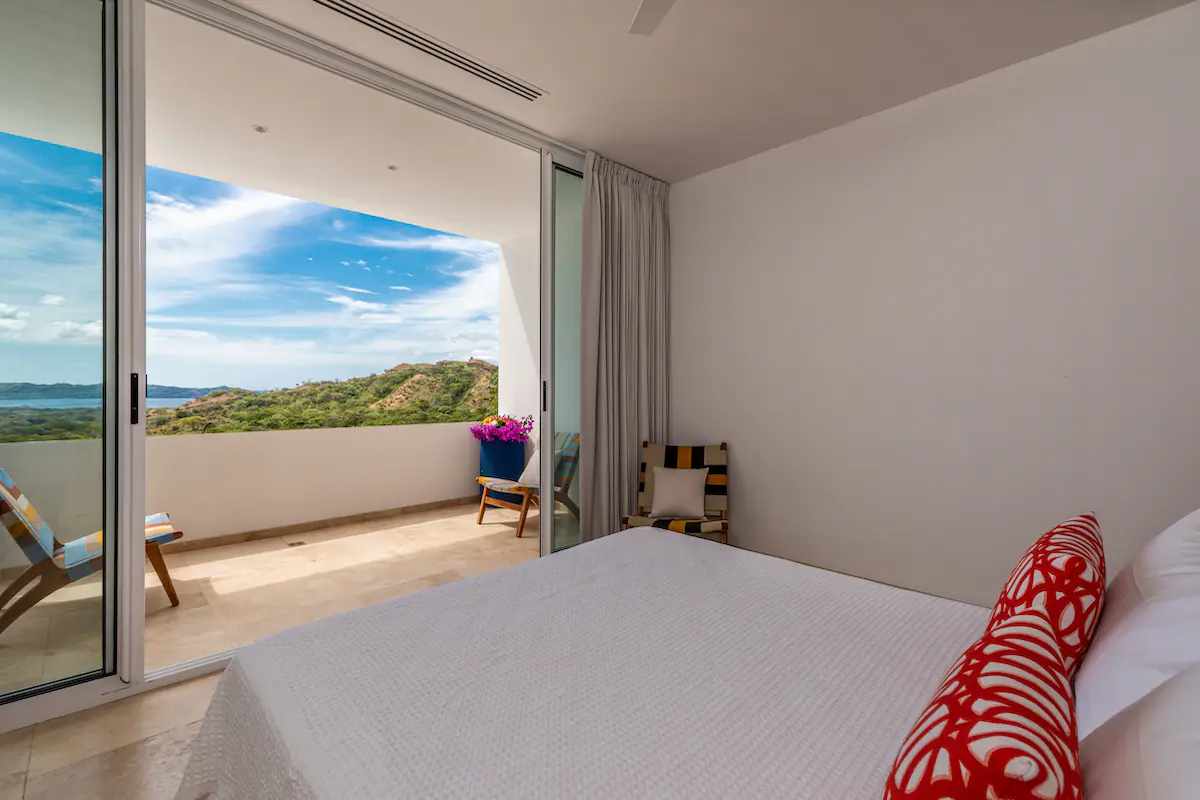 Chambre avec balcon ouvert donnant sur un paysage tropical et l'océan ; lit, chaises, murs blancs.