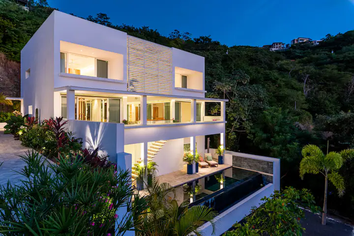 Maison blanche moderne avec piscine à flanc de colline, au crépuscule. Une végétation luxuriante entoure le bâtiment.