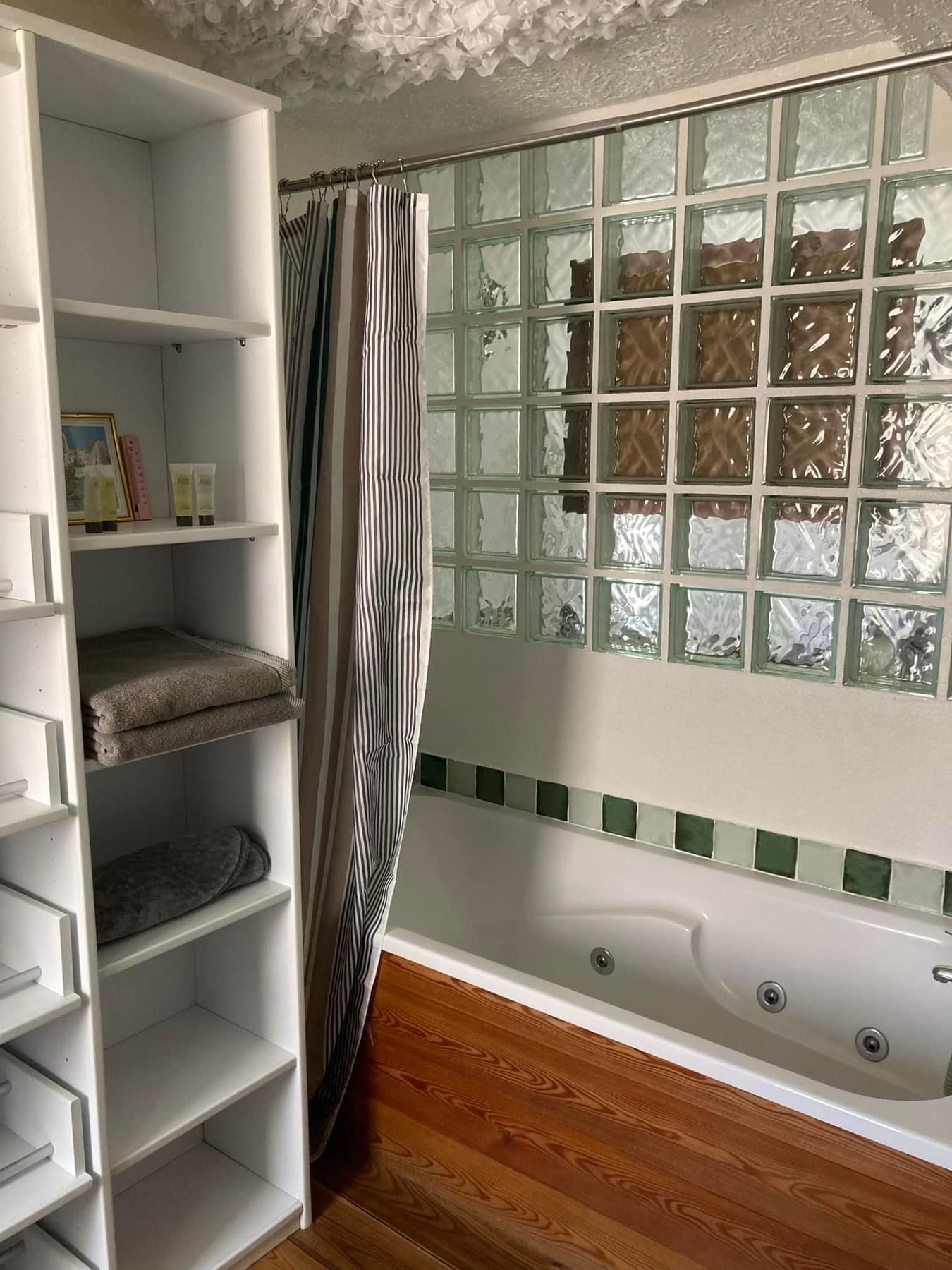Salle de bain avec étagères blanches, baignoire, mur en blocs de verre et parquet.