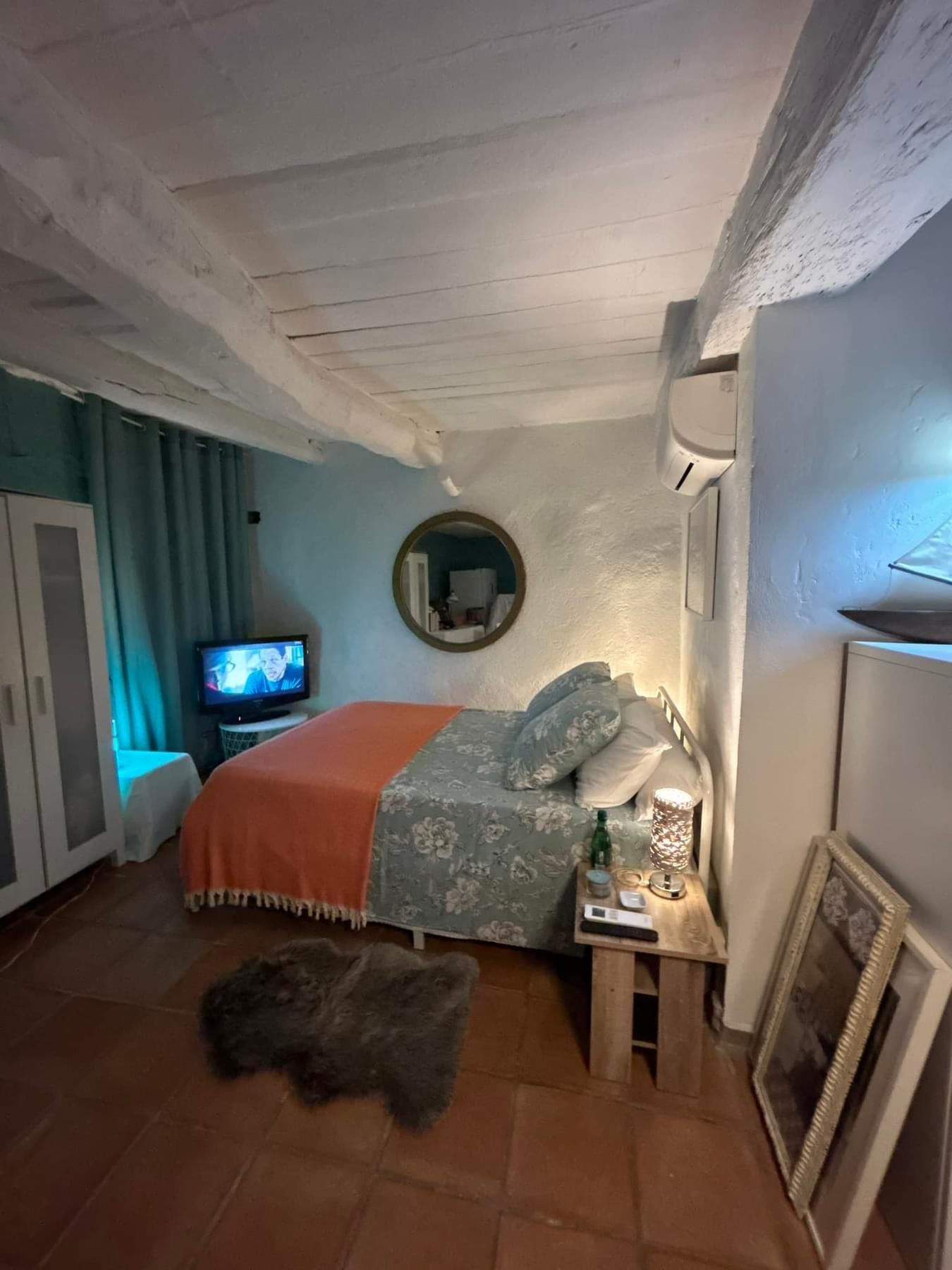 Chambre avec lit, rideaux bleu sarcelle, armoire blanche et couverture orange ; sol en carrelage marron.