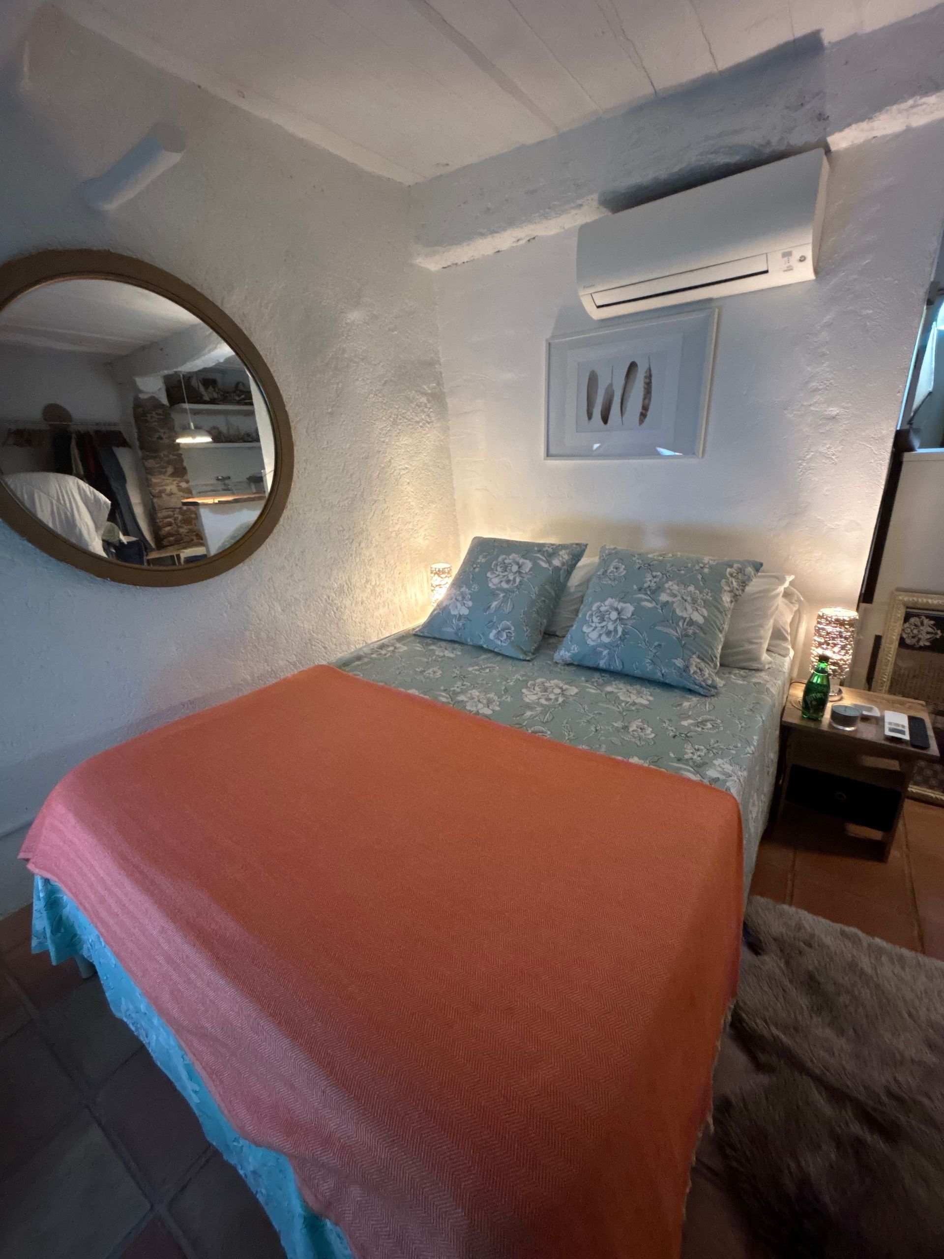 Chambre avec un lit, un miroir rond et un climatiseur. Couverture couleur pêche, linge de lit fleuri et table de chevet.