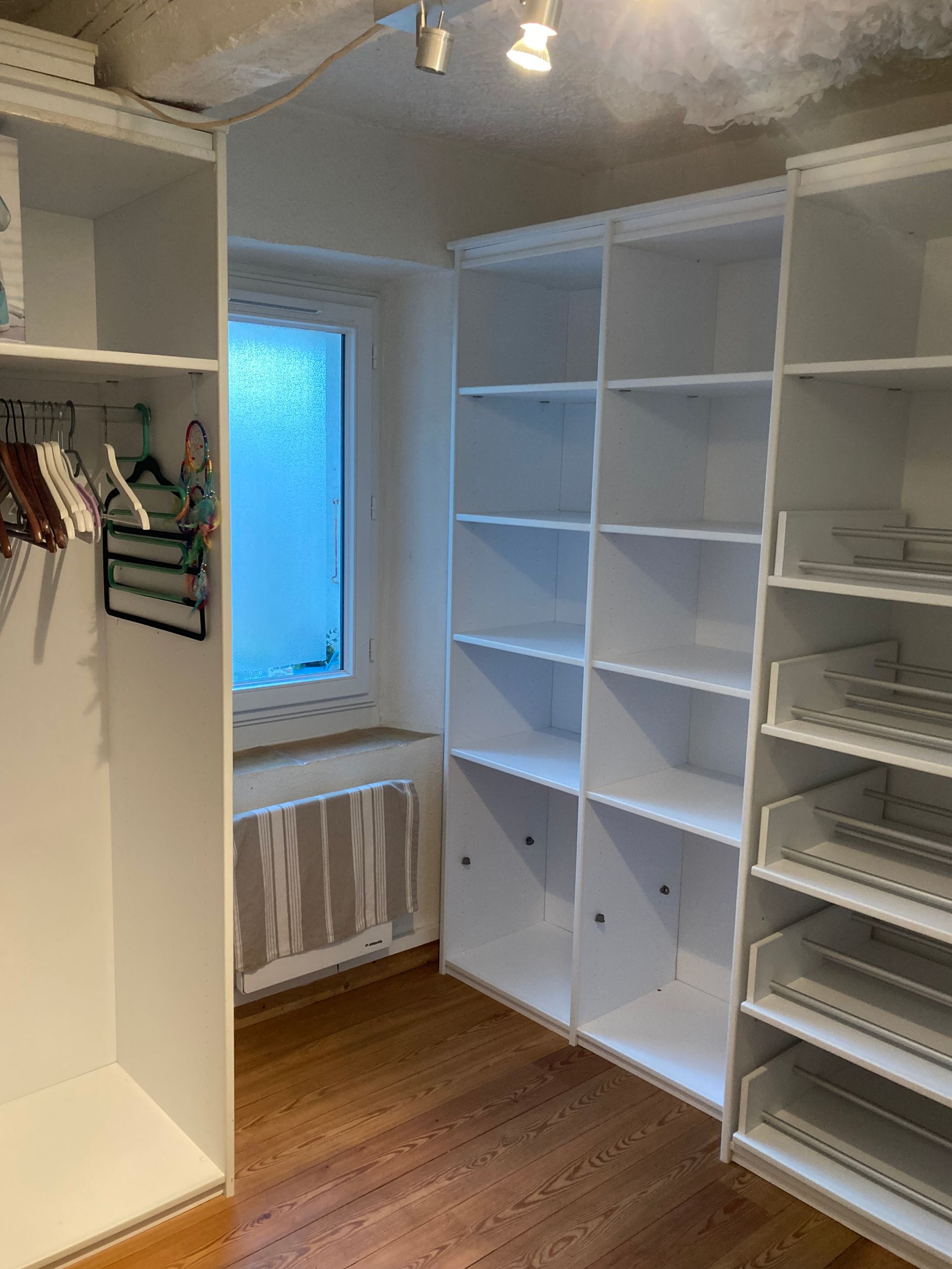 Placard blanc avec étagères et tiroirs dans une pièce avec une petite fenêtre et du parquet.