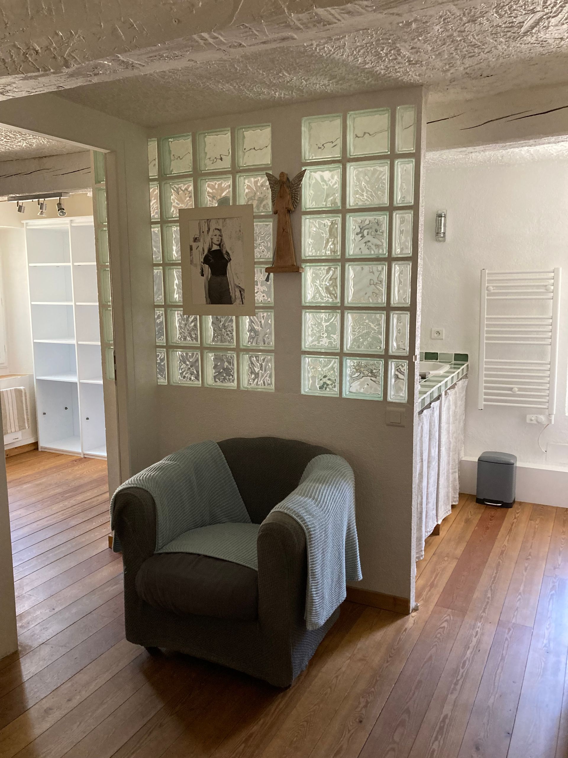 Intérieur cosy avec fauteuil, mur en briques de verre, photo et vue sur une autre pièce avec armoires blanches.