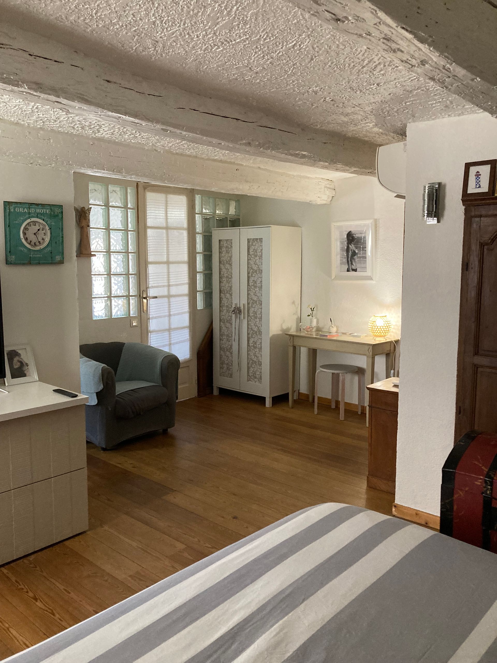 Chambre avec parquet, poutres blanches et meubles anciens : fauteuil, armoire, bureau et malle.