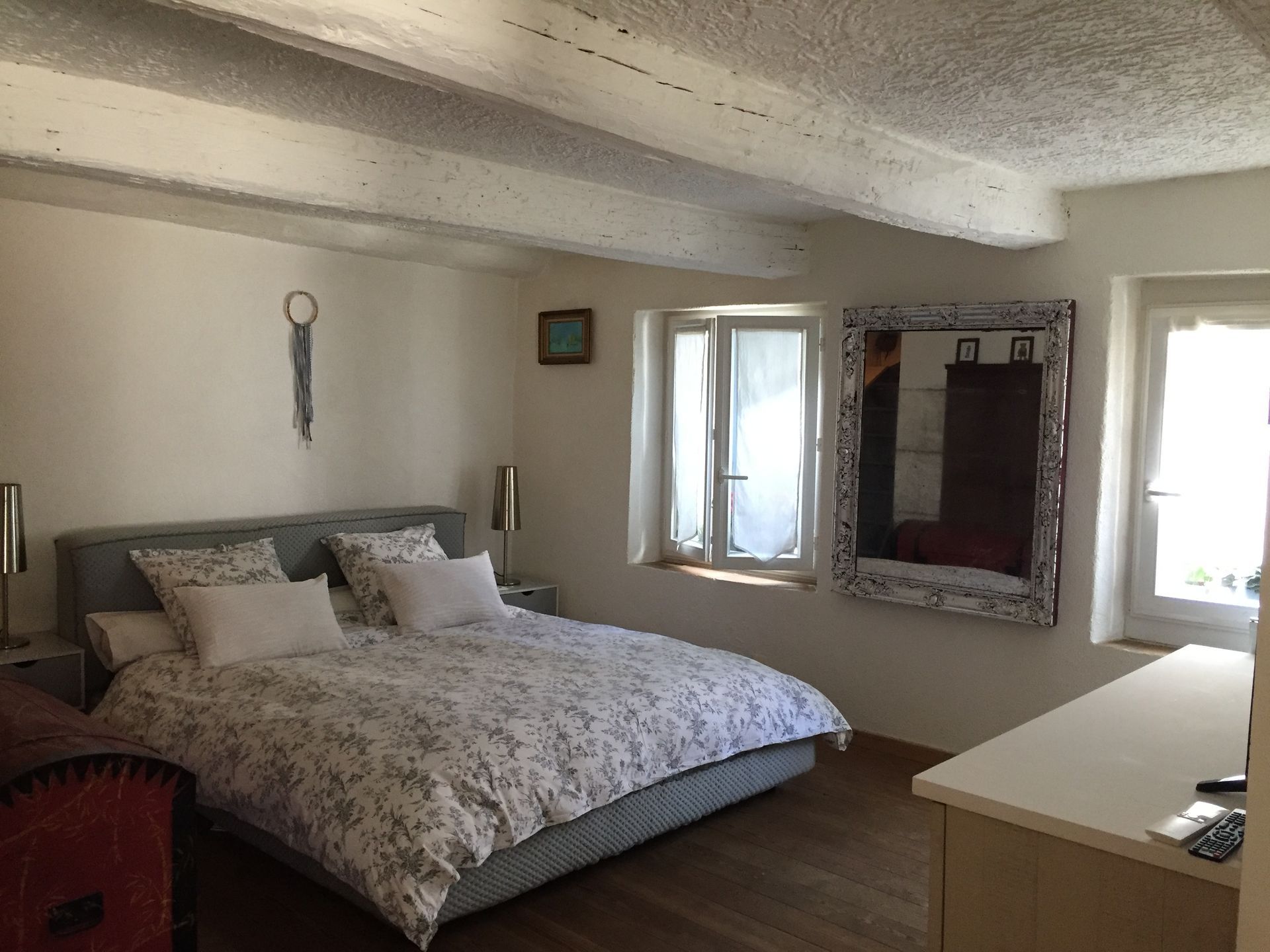 Chambre avec lit, fenêtres, miroir, murs blancs et poutres en bois au plafond.