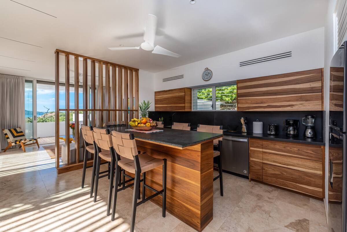 Cuisine moderne avec armoires en bois, îlot central et tabourets de bar. Ouverte sur une terrasse avec vue sur l'océan.