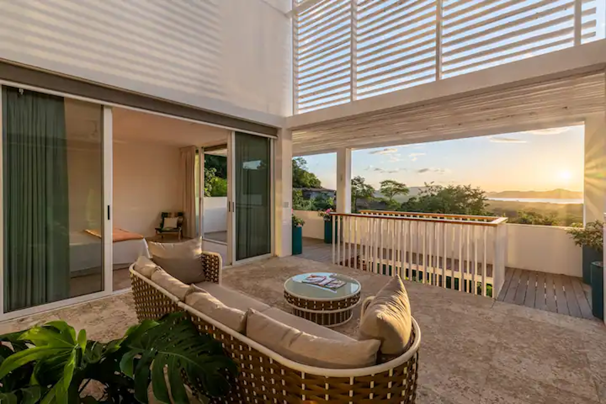 Terrasse extérieure avec canapé en osier, table basse ronde et portes coulissantes en verre menant à une chambre.