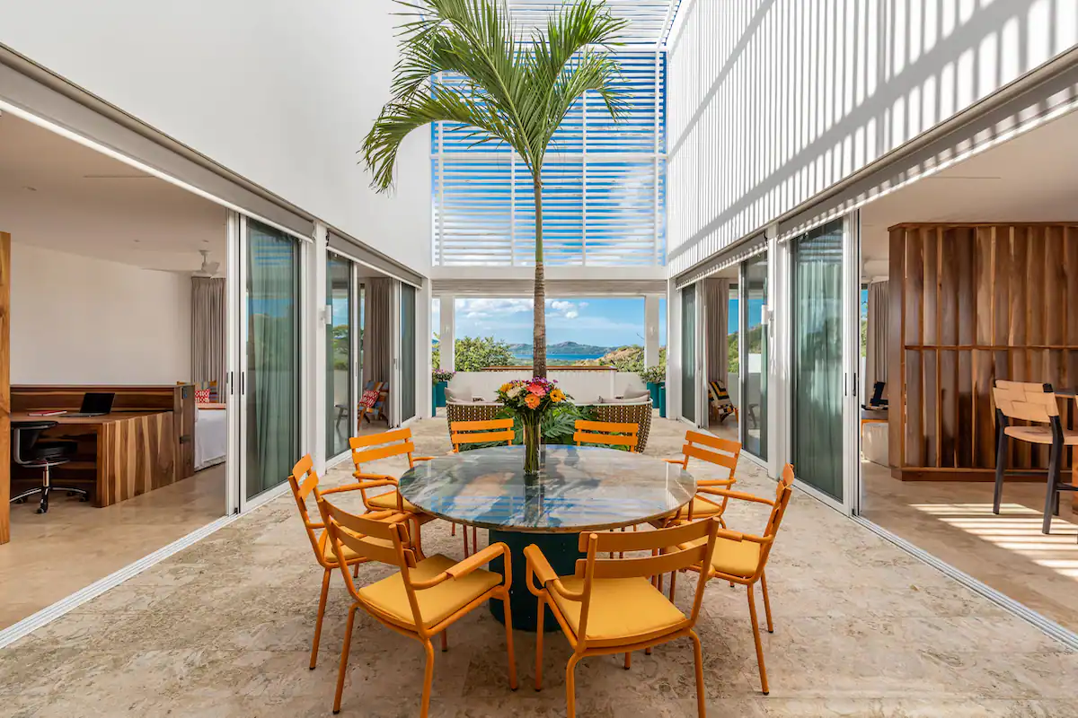 Un espace repas extérieur avec une table ronde, des chaises jaunes et un palmier, flanqué de pièces avec des portes vitrées.
