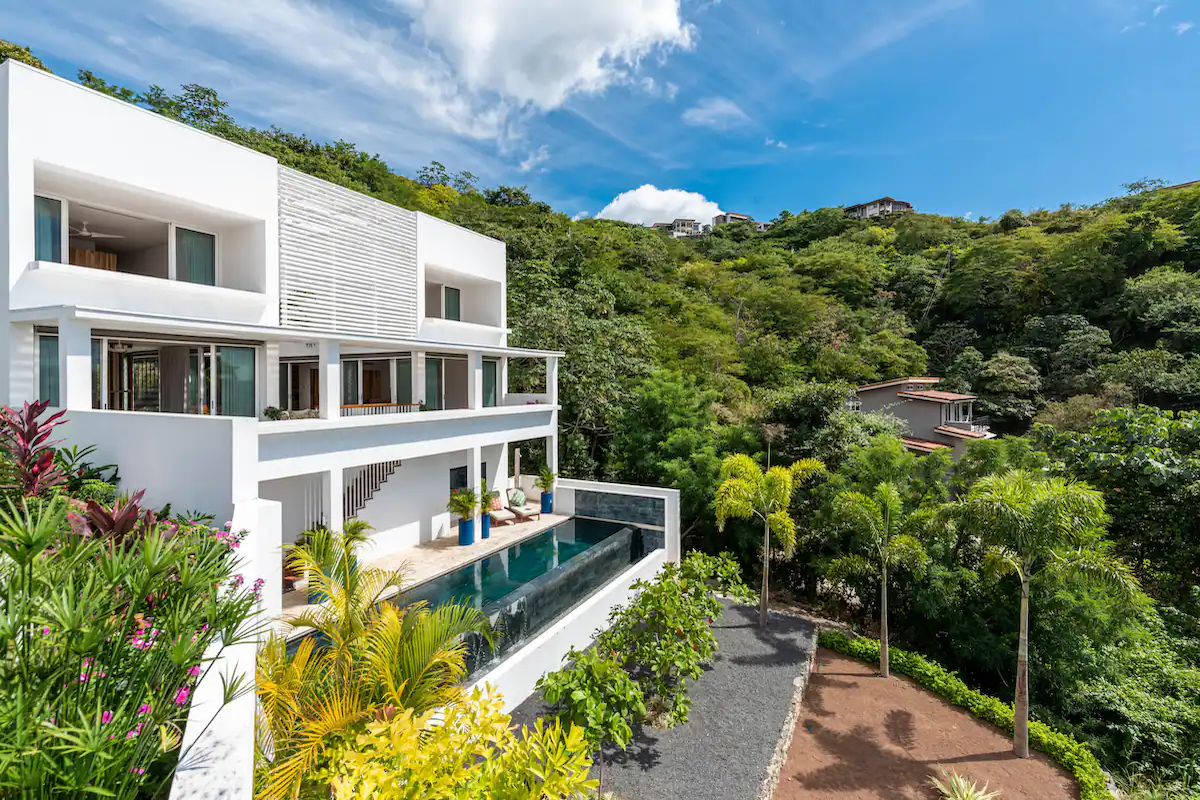 Maison blanche moderne avec piscine, entourée d'une végétation luxuriante, à flanc de colline.