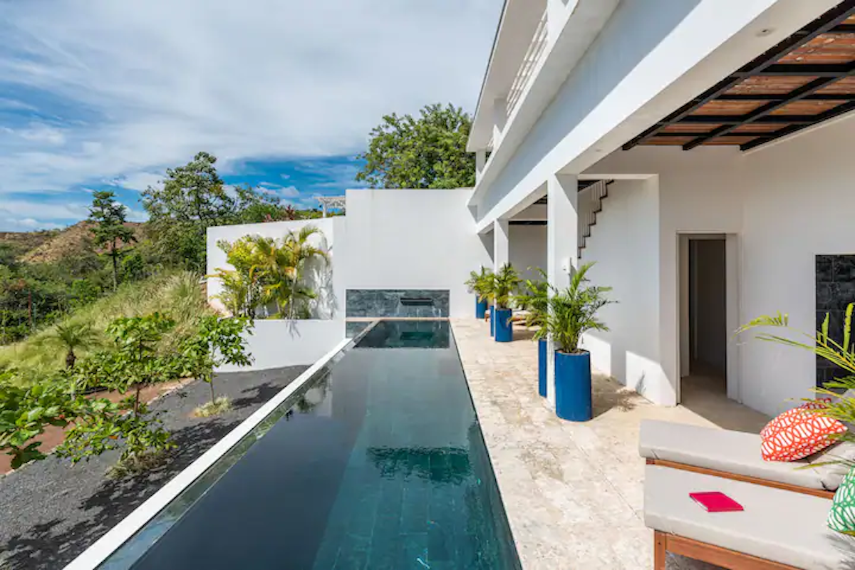 Longue et étroite piscine à côté d'une maison blanche. Des pots bleus garnis de plantes bordent l'allée.