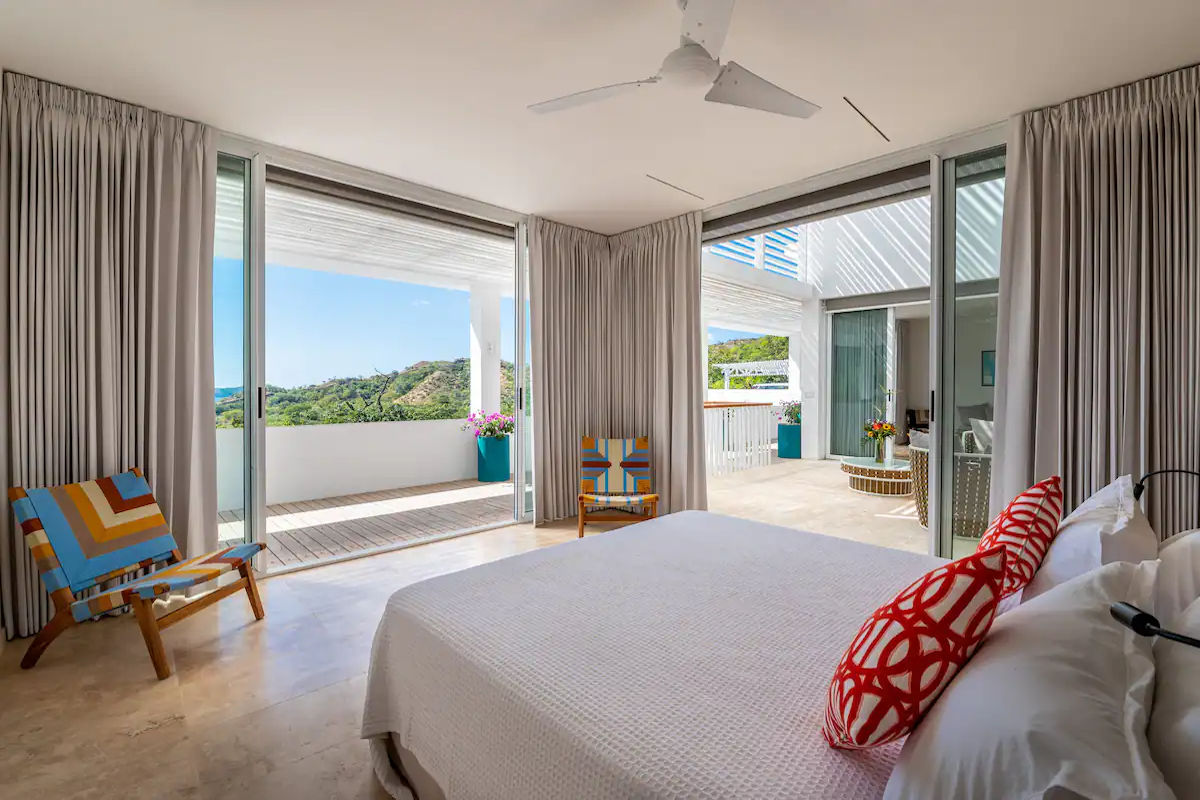 Chambre lumineuse avec un lit blanc, des portes ouvertes sur un balcon et des vues panoramiques.