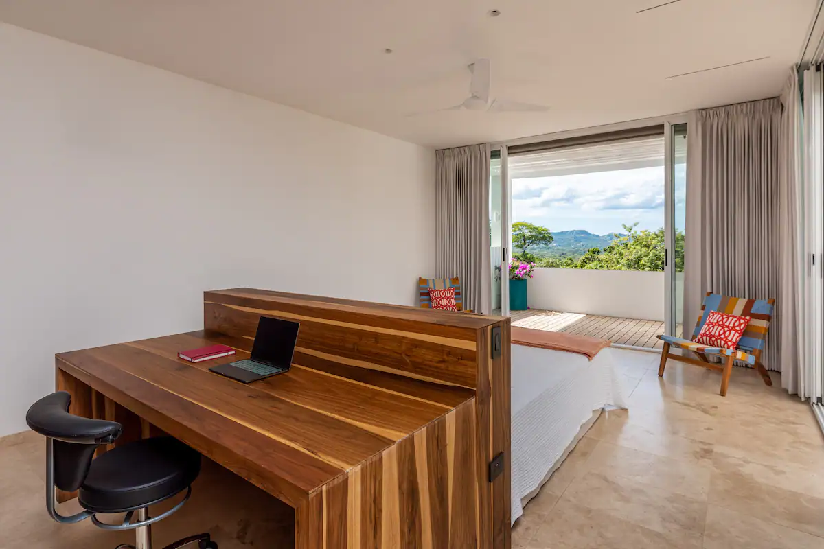 Chambre avec bureau en bois, lit et balcon donnant sur les arbres.