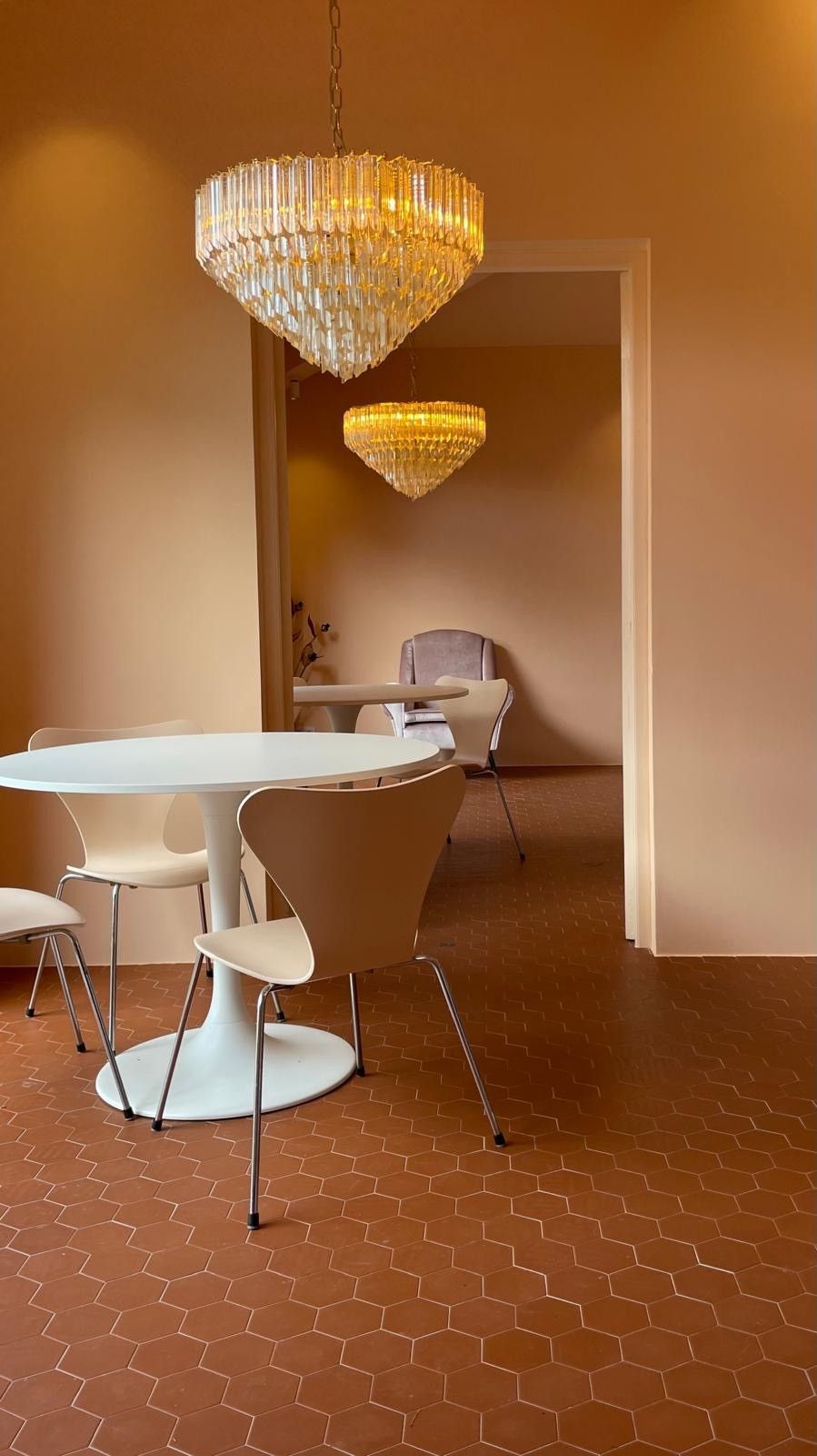 Salle à manger avec table tulipe blanche, chaises, lustres en cristal et sol brun rougeâtre.