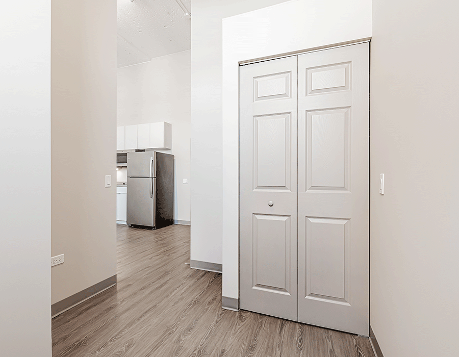 A hallway with a closet and a refrigerator in the background.