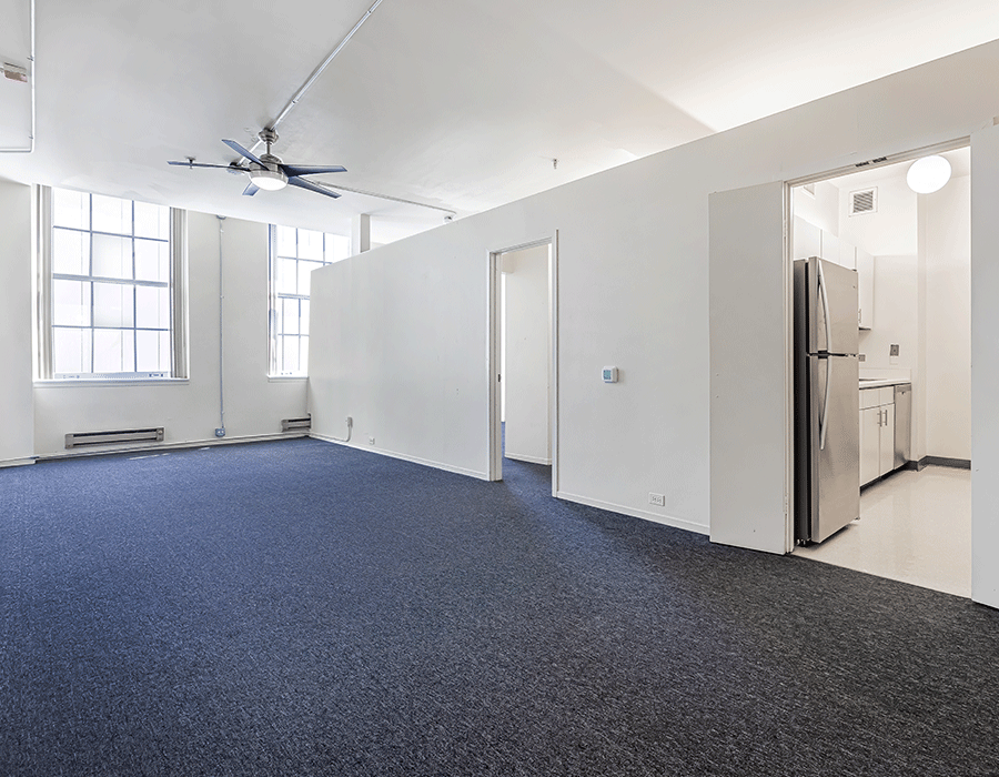 A large empty room with a ceiling fan and a refrigerator.