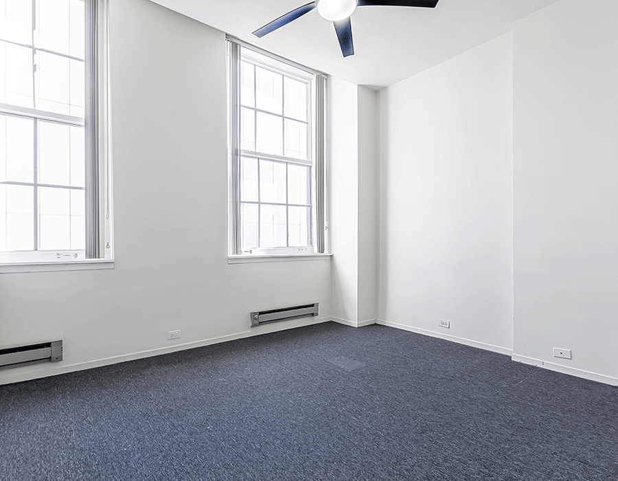 An empty room with a ceiling fan and two windows.