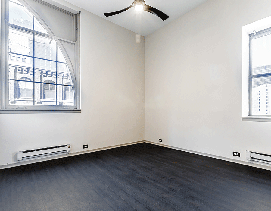 An empty room with a ceiling fan and two windows.