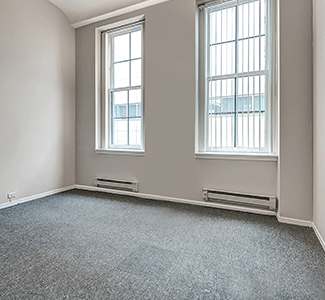 An empty room with two windows and a gray carpet.