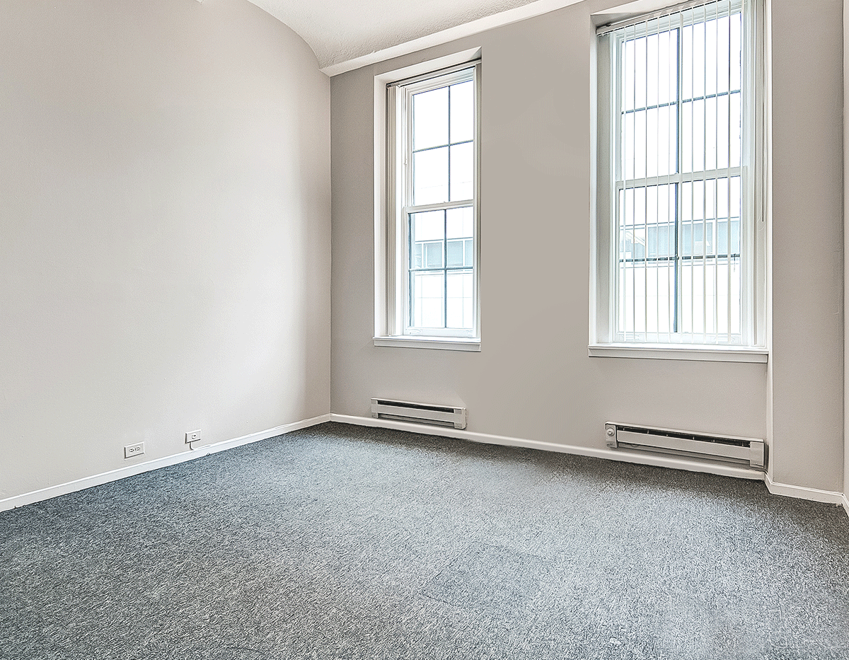 An empty room with a carpeted floor and two windows.