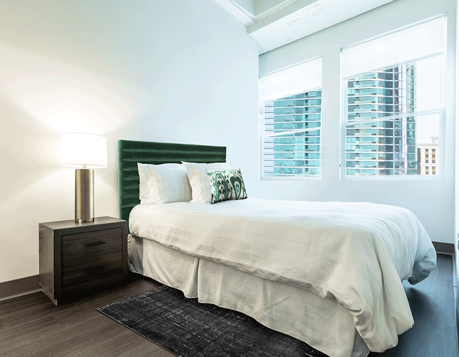 A bedroom with a bed , nightstand , lamp and window.