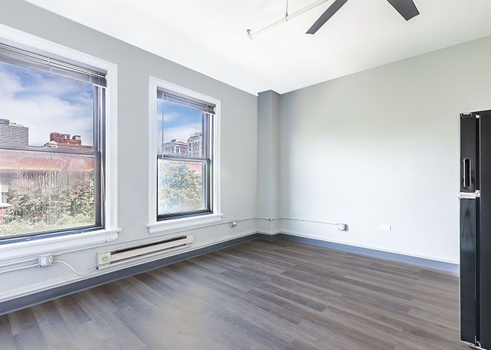 An empty living room with hardwood floors , two windows , a refrigerator and a ceiling fan.