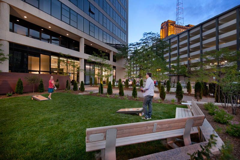 Two men are playing cornhole in a park in front of a building.