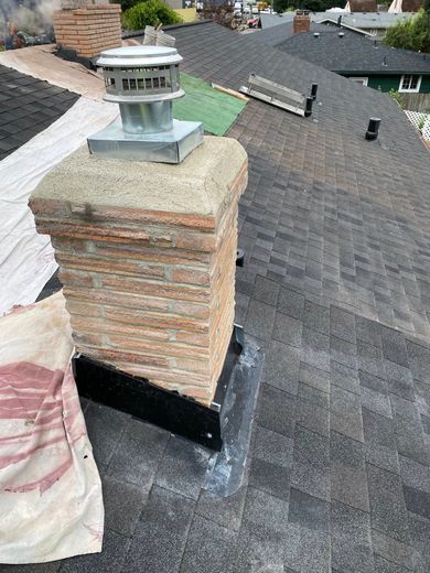 Chimney on a dark shingled roof, topped with a metal cap, near other rooftops.