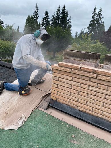 Person cutting brick chimney on a rooftop, wearing protective gear, surrounded by dust.