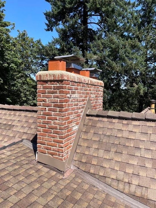 Brick chimney on a brown shingled roof with three metal chimney caps, set against trees and a blue sky.
