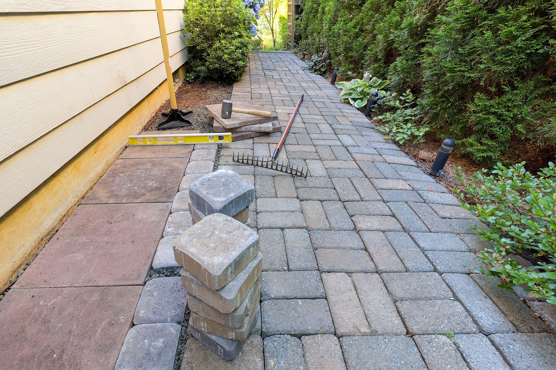 Pathway construction: Brick pathway being built along a building and garden with tools and stacked bricks.