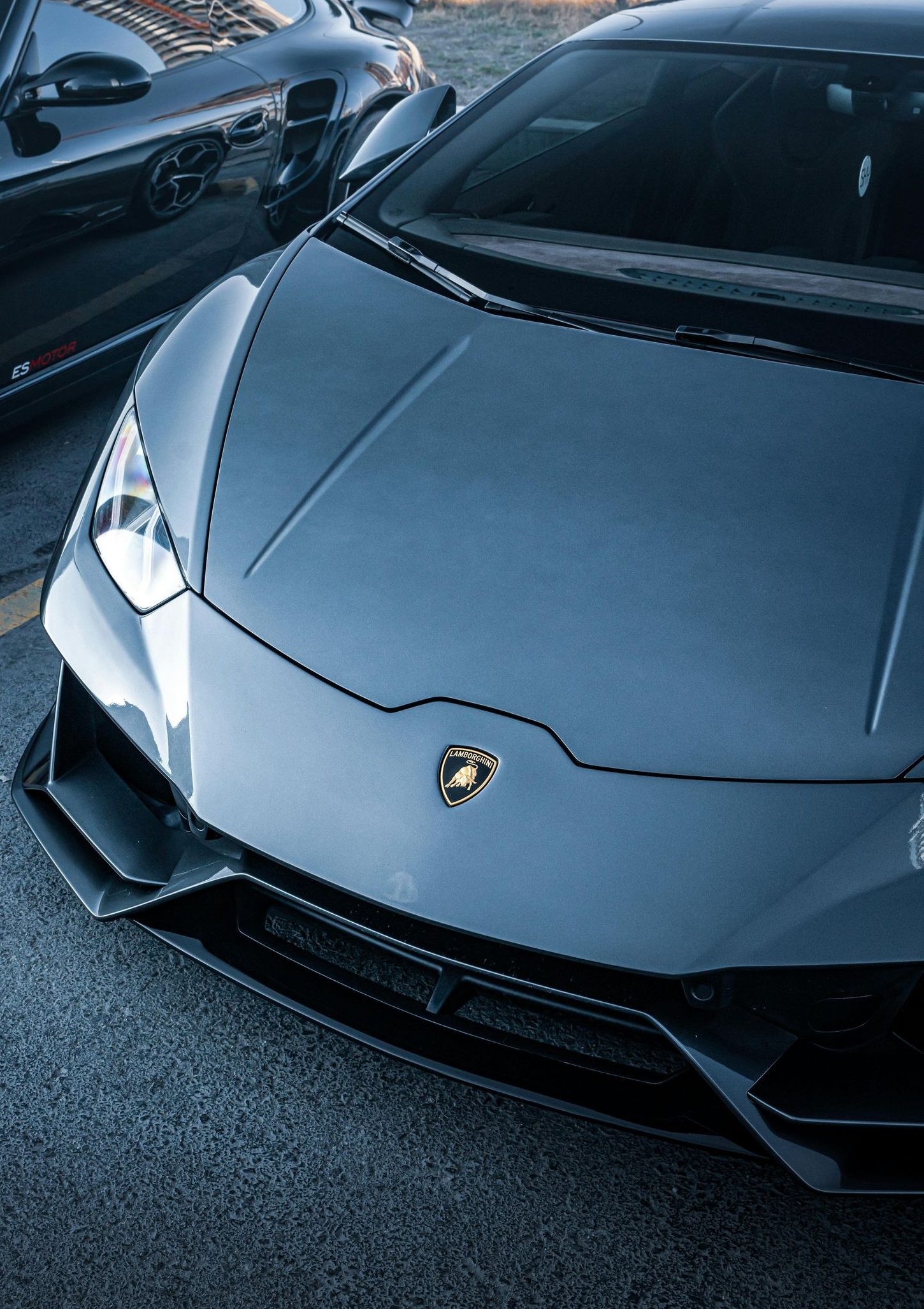 Lamborghini and Porsche sports cars parked in an asphalt lot, dusk lighting.
