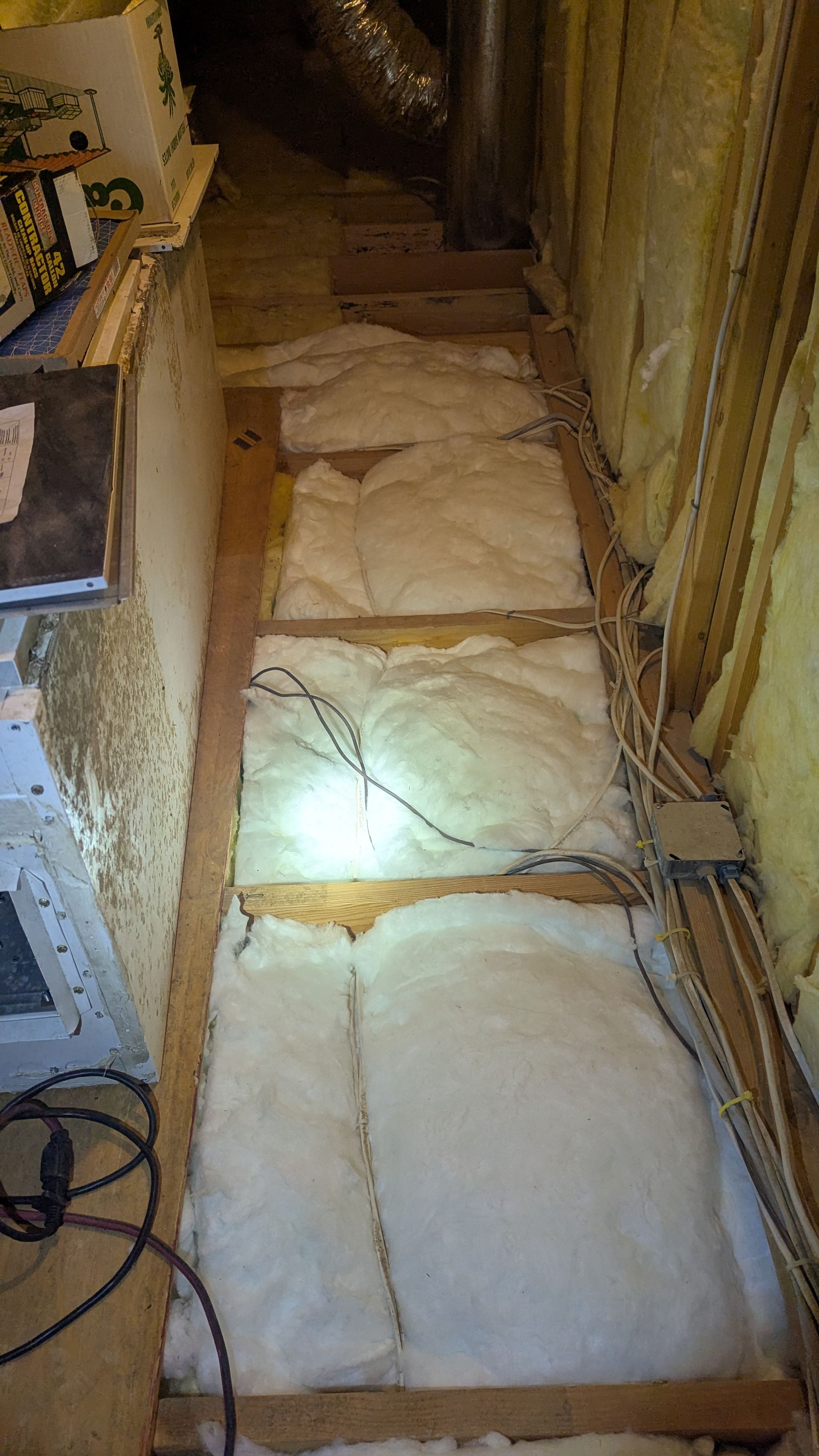 Attic with exposed insulation, wooden beams, and electrical wiring. A small walkway is present.