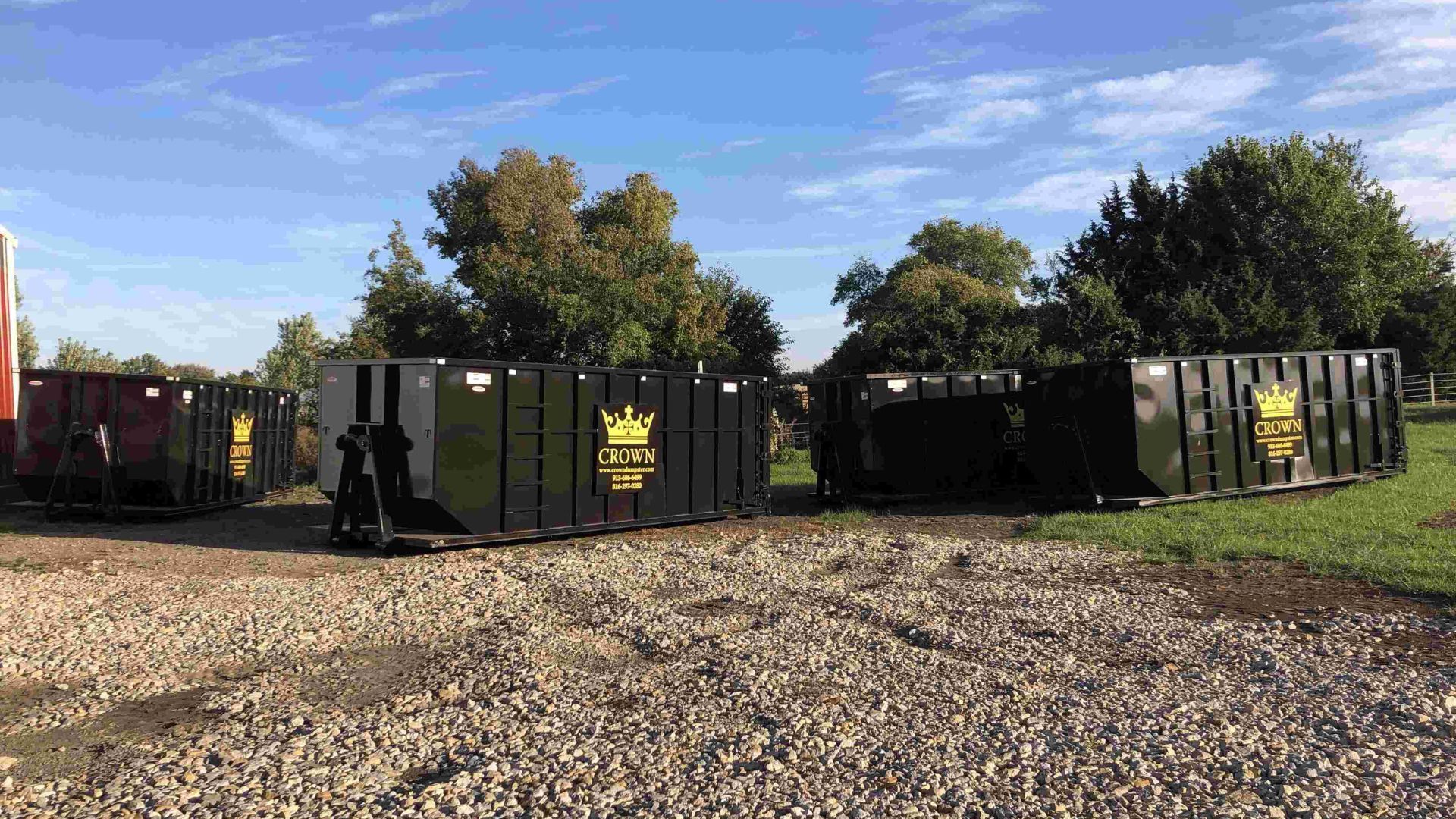 Large black dumpsters lined up on a gravel lot under a blue sky, with trees in the background