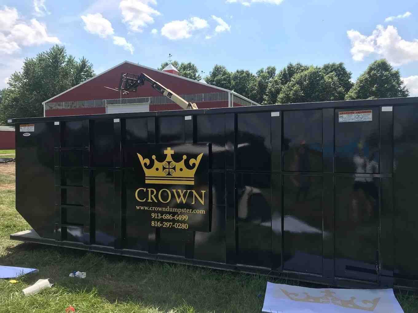 Black dumpster with gold crown logo in front of a red barn.