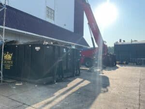 Red crane extending towards a building with a black dumpster in front. Sunlight.