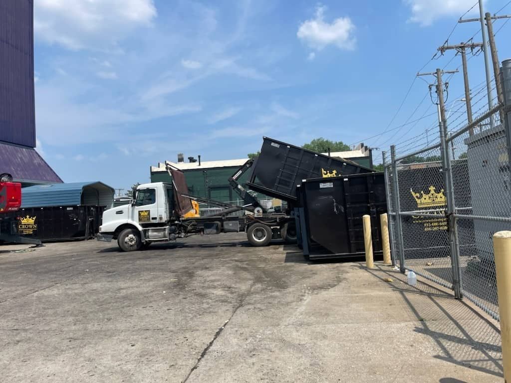 Dump truck unloading a black dumpster on a paved lot, beside a chain-link fence and a building.