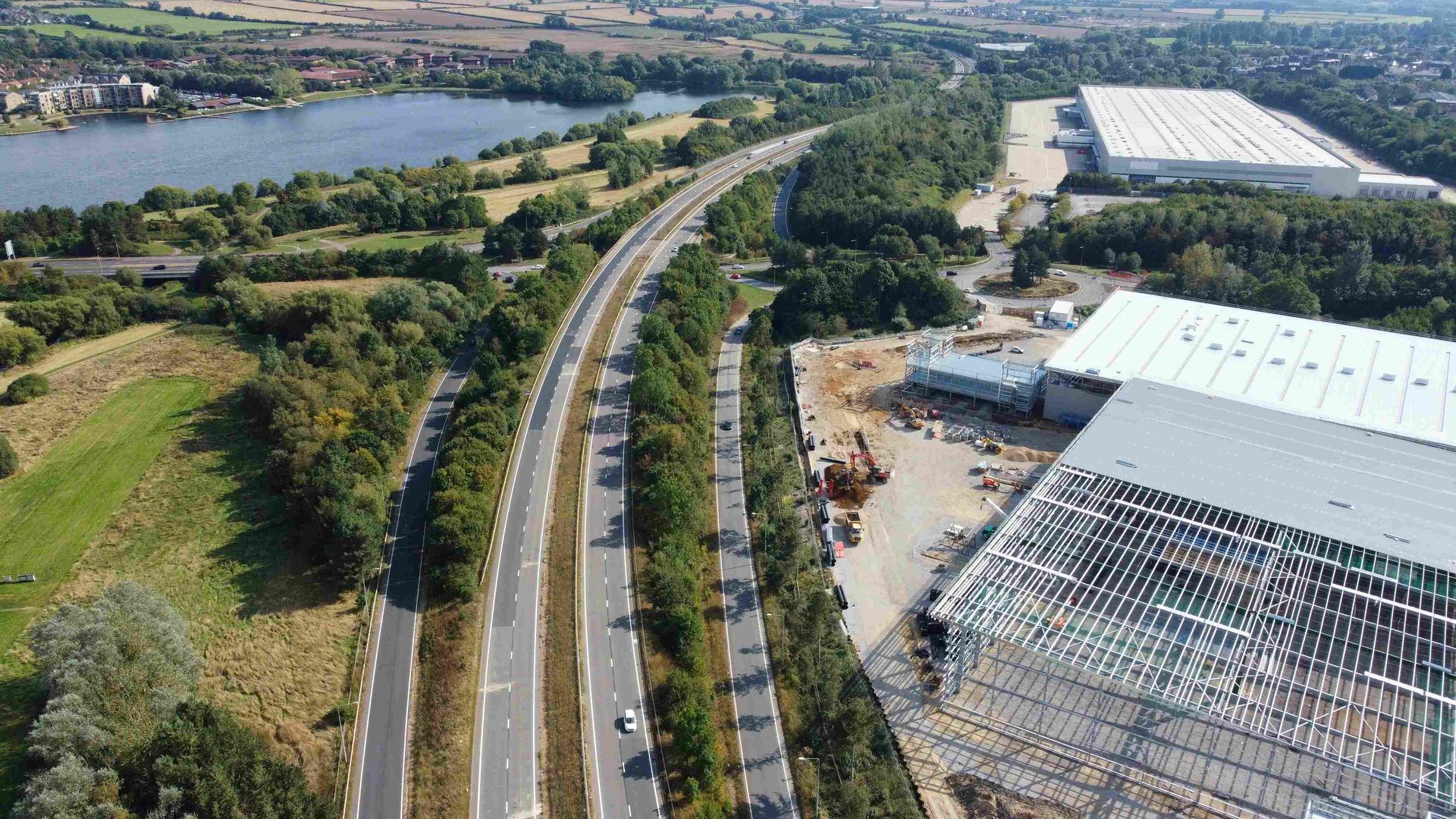 Aerial view of a highway with traffic, a lake, trees, and buildings under construction.
