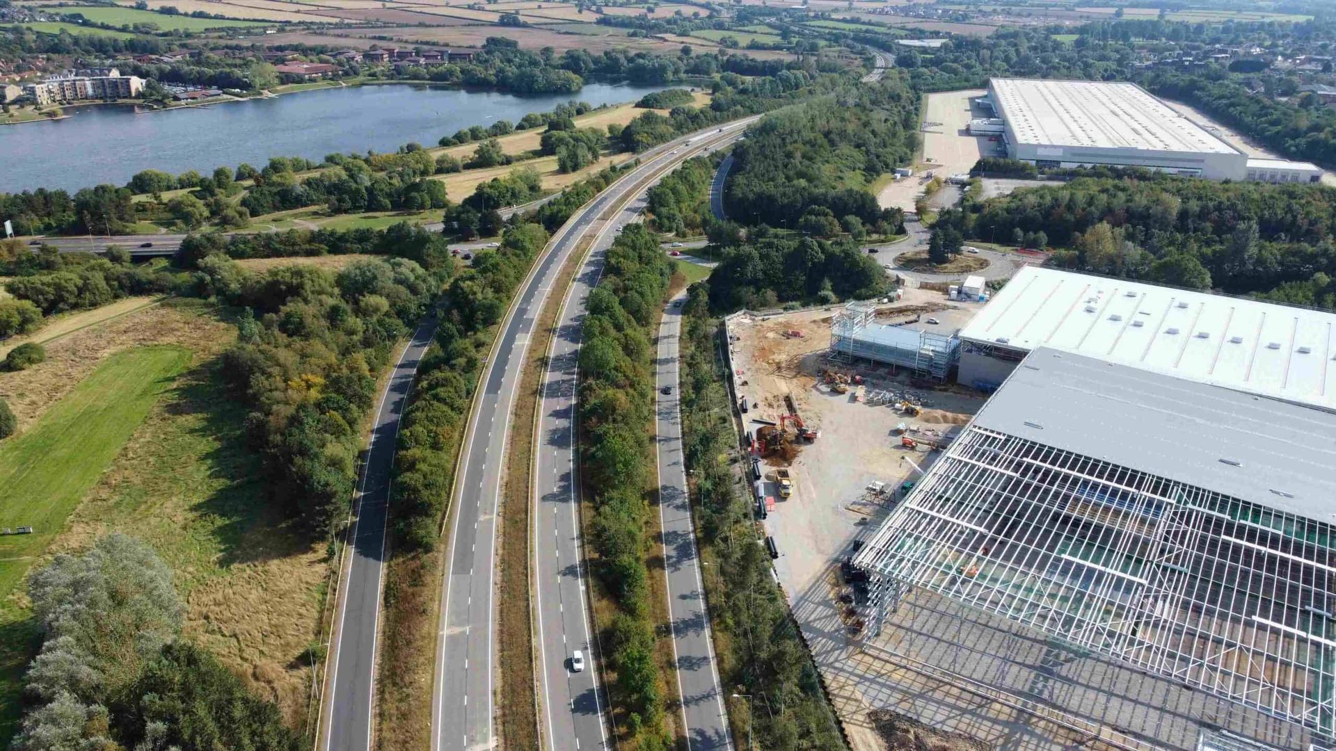 Aerial view of a highway with traffic, a lake, and a large warehouse under construction on a sunny day.
